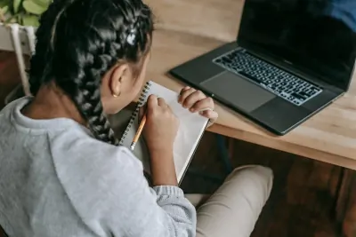 A young woman writing in a notebook beside a laptop